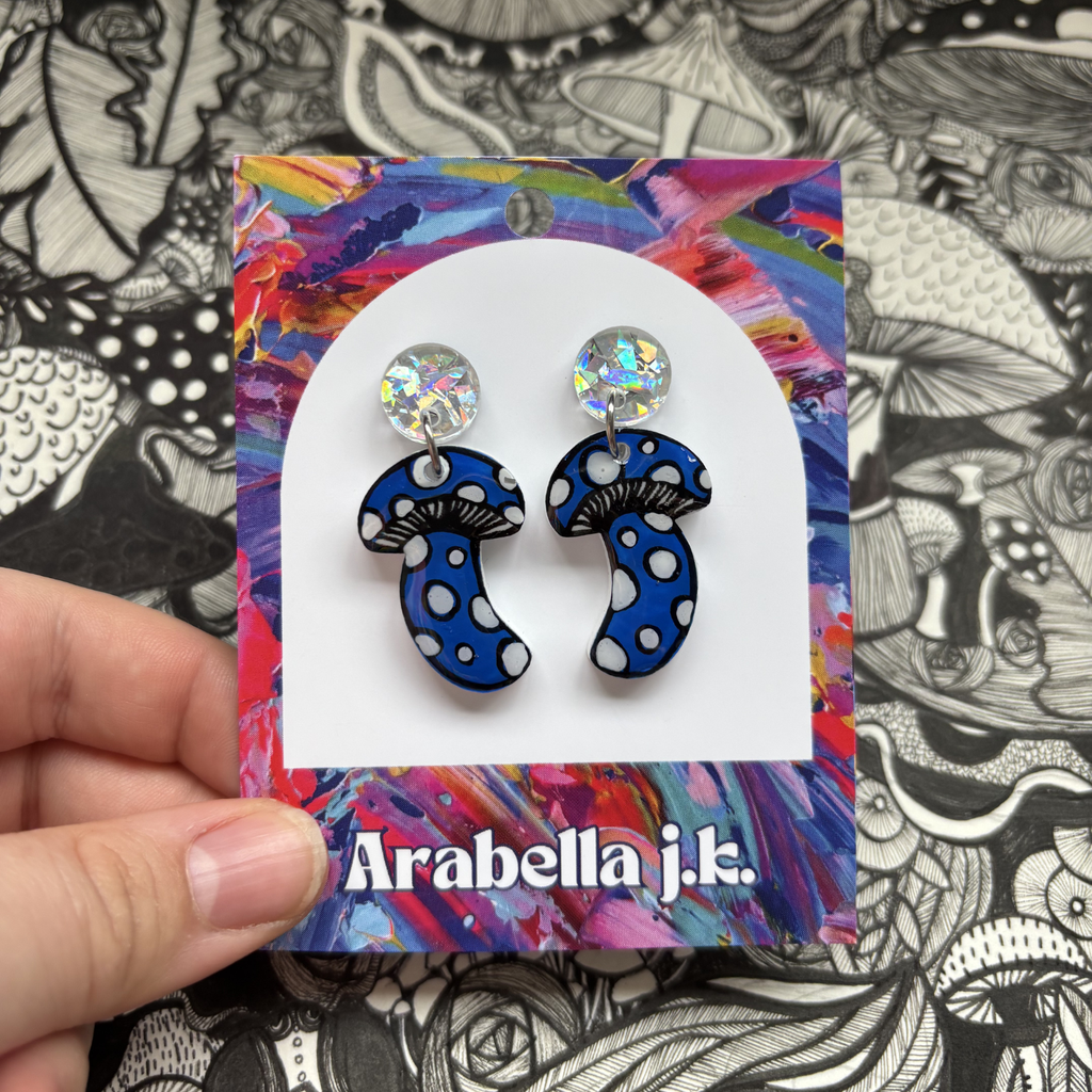 Blue and white polka dot earrings on a colorful 'Arabella j.k.' branded card against a black and white patterned background.