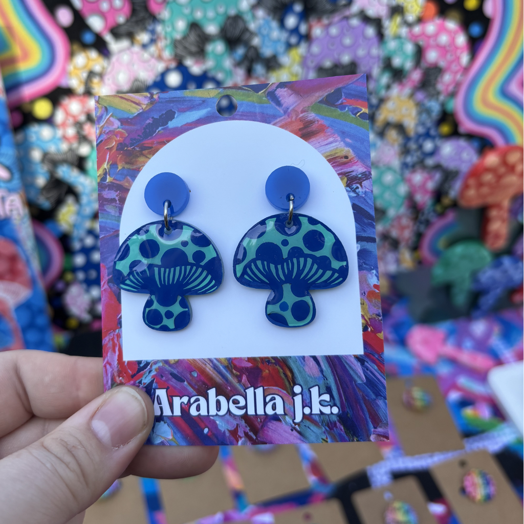 Colorful mushroom-shaped earrings on a 'Arabella j.k.' branded card against a multicolored background.