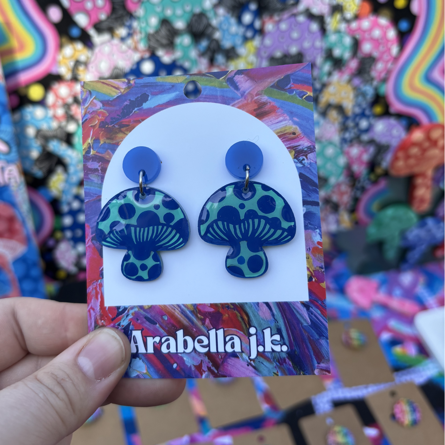 Colorful mushroom-shaped earrings on a 'Arabella j.k.' branded card against a multicolored background.