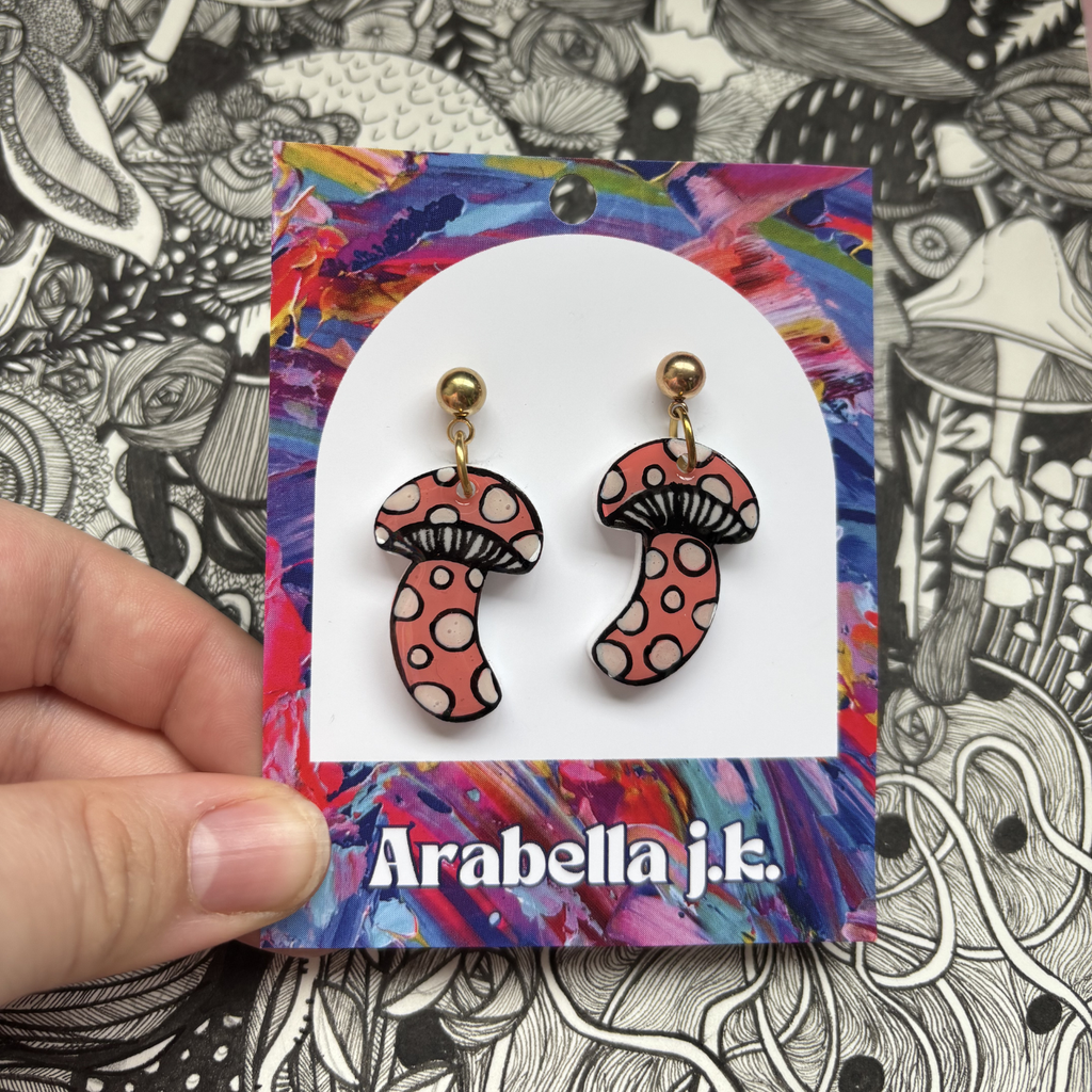 Mushroom-shaped earrings on a colorful 'Arabella j.k.' card against a black and white patterned background.