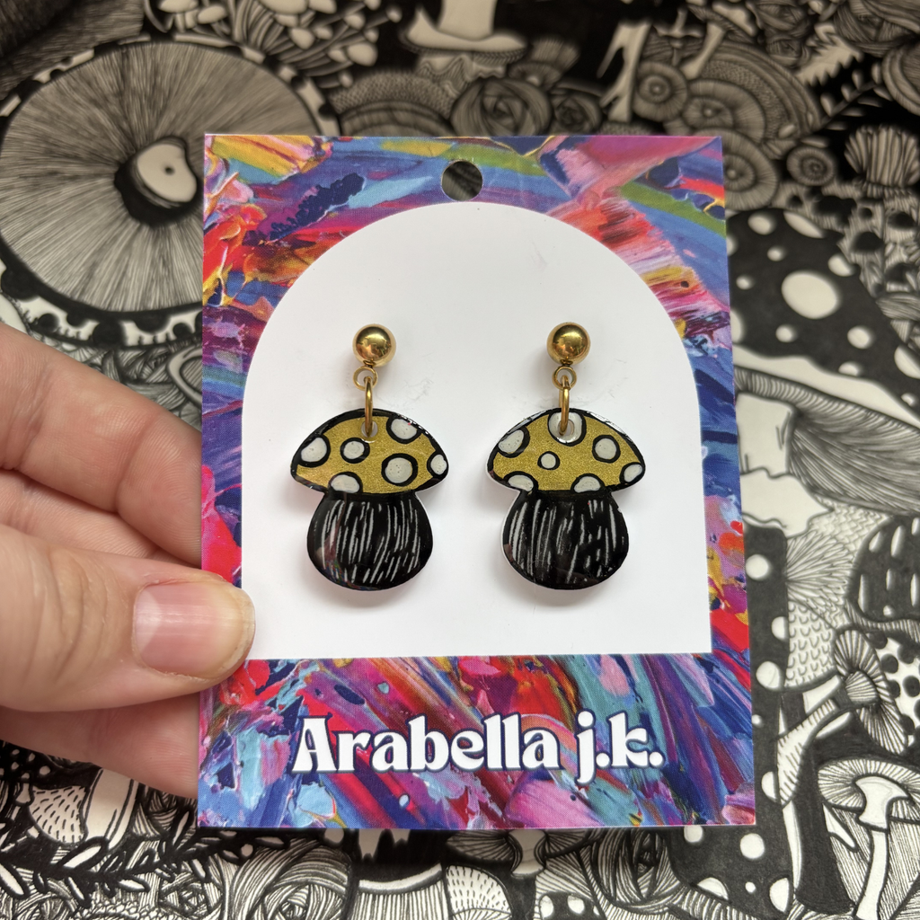 Mushroom-shaped earrings on a 'Arabella j.k.' branded card against a black and white patterned background.