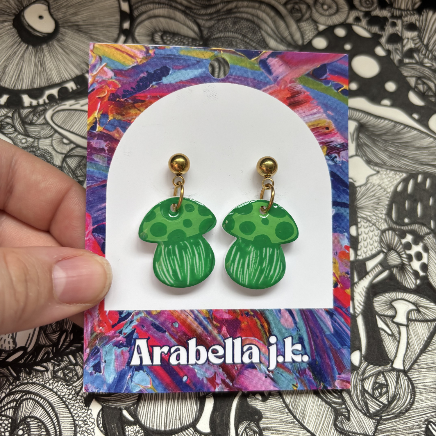 Green mushroom earrings on a colorful 'Arabella j.k.' card against a black and white patterned background.