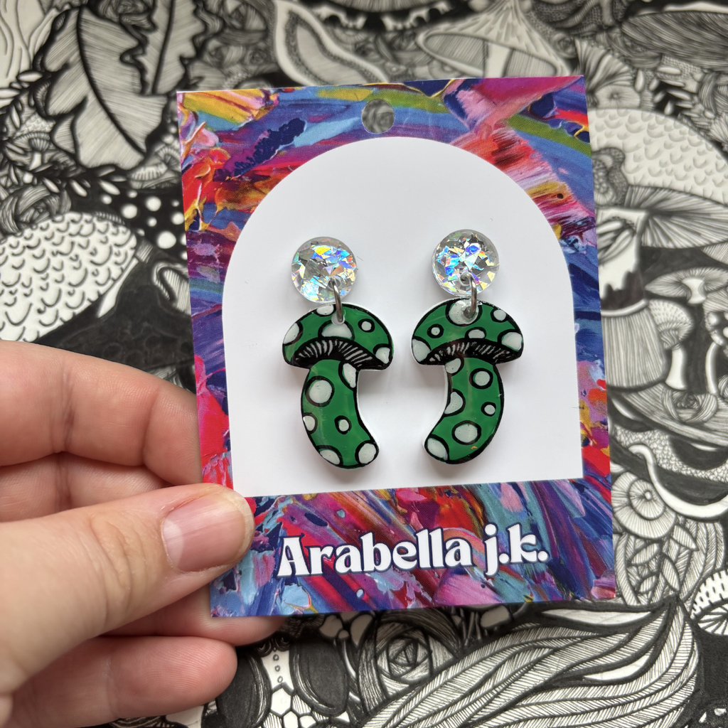 Green polka dot mushroom earrings on a colorful 'Arabella j.k.' card