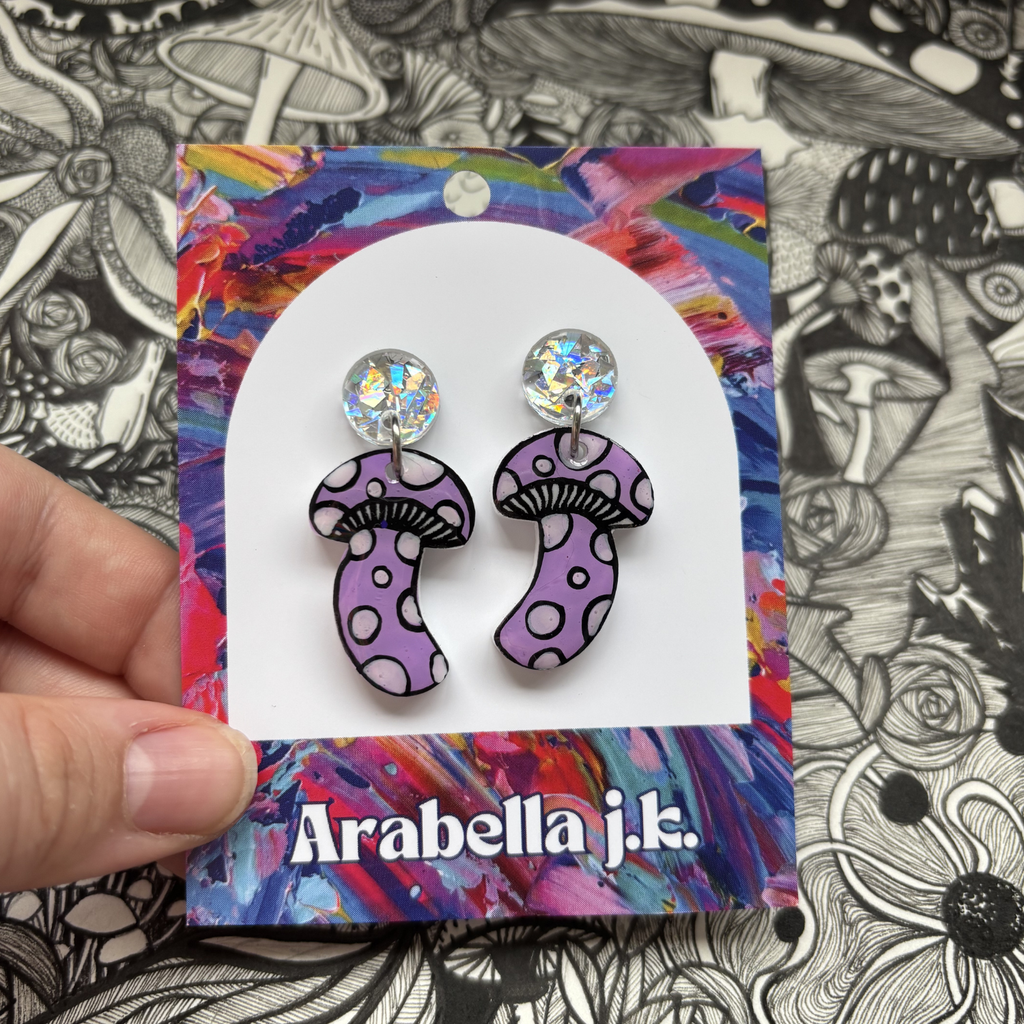 Purple mushroom earrings on a colorful 'Arabella j.k.' card against a black and white floral background.