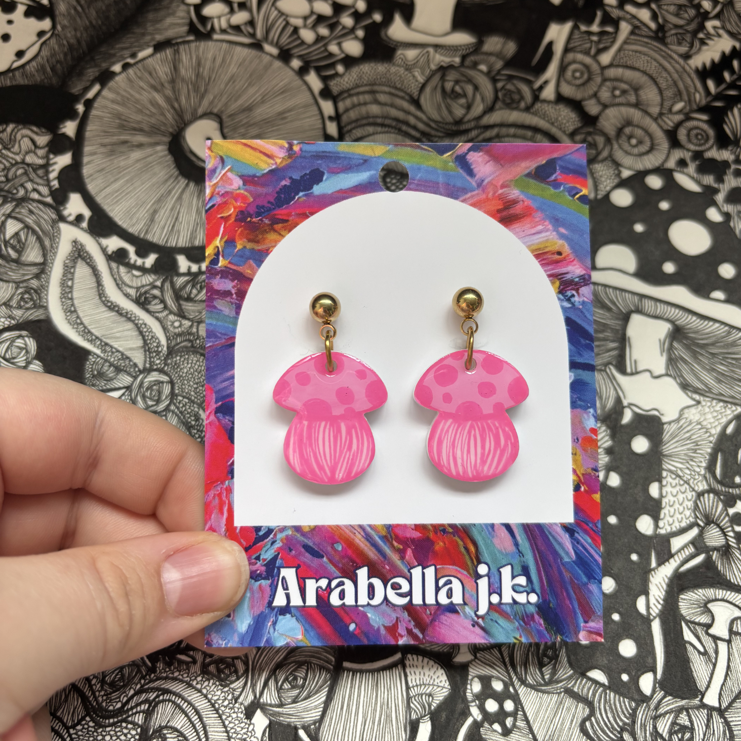 Pink mushroom-shaped earrings on a colorful 'Arabella j.k.' card against a black and white patterned background.