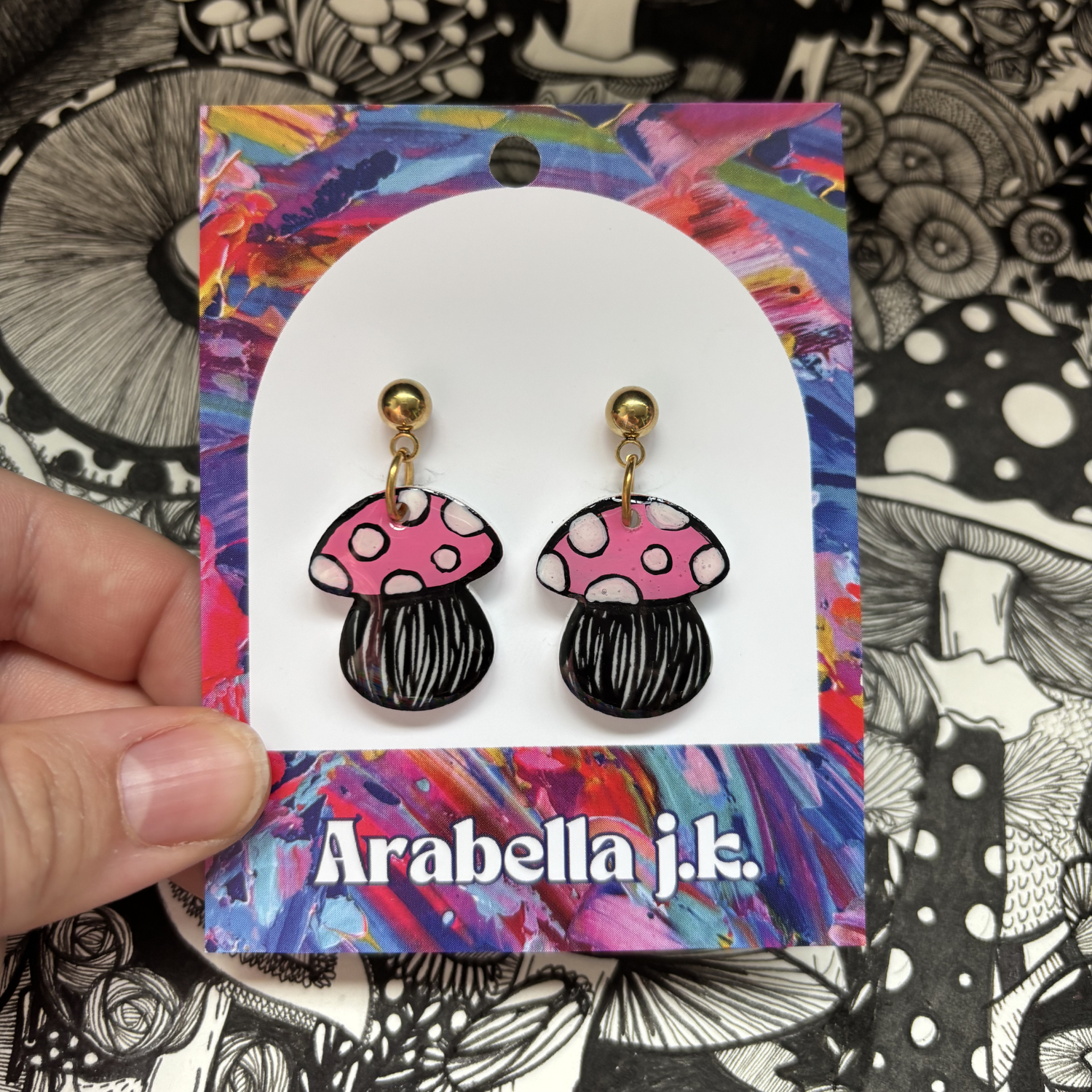 Mushroom-shaped earrings on a colorful 'Arabella j.k.' card against a patterned background