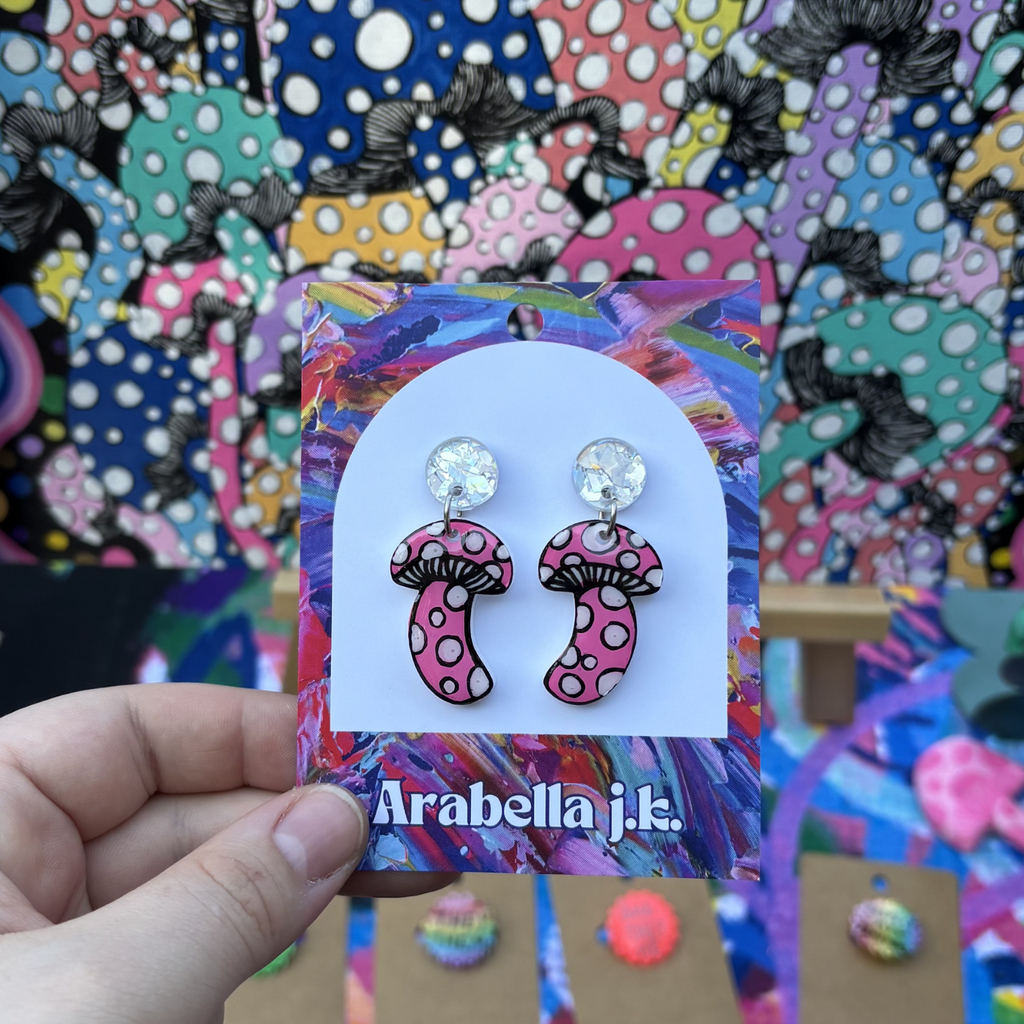 Polka dot mushroom earrings on a colorful 'Arabella j.k.' card held against a vibrant background.