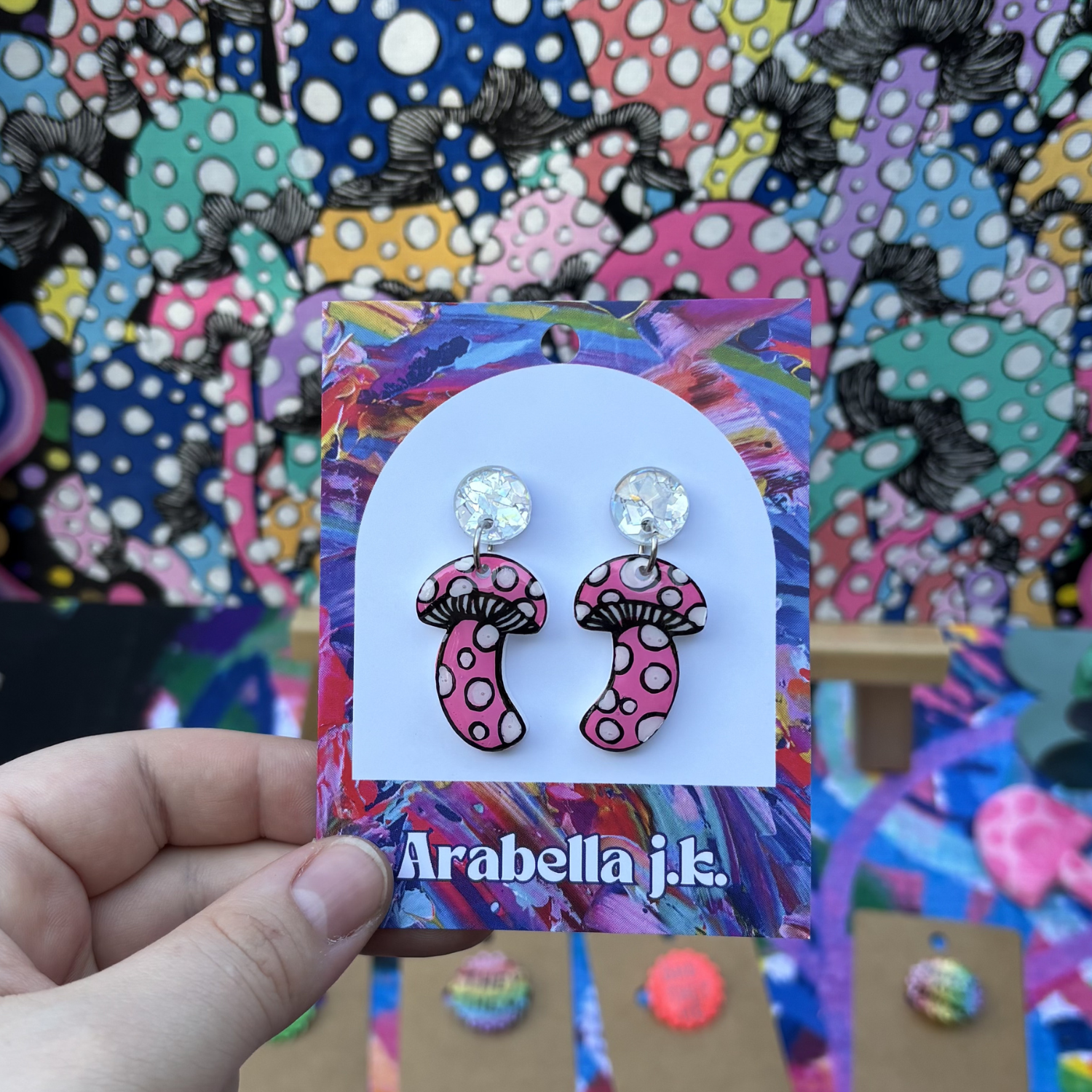 Polka dot mushroom earrings on a colorful 'Arabella j.k.' card held against a vibrant background.