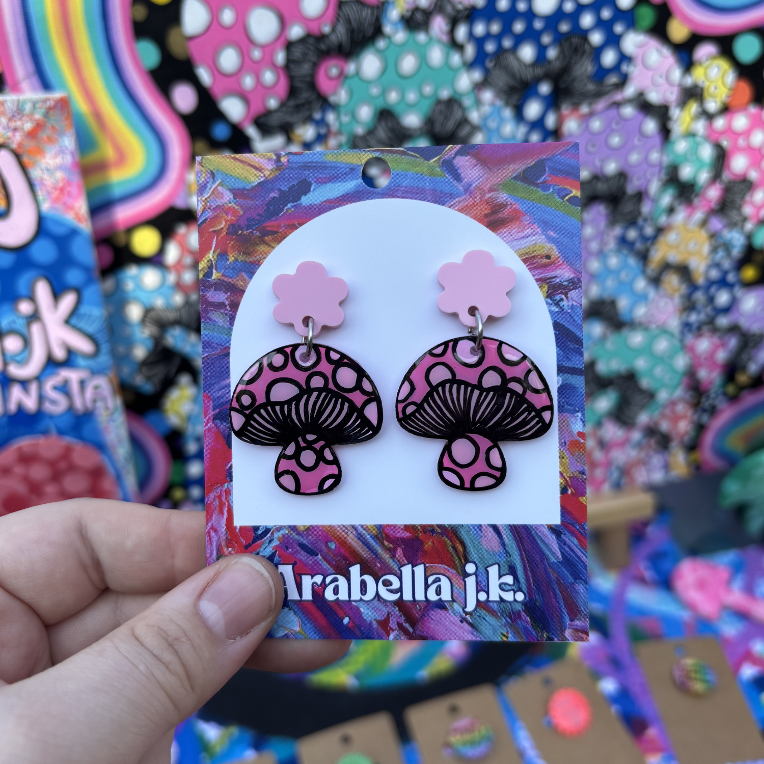 Pink and black mushroom-shaped earrings on a colorful 'Arabella j.k.' card held by a hand.