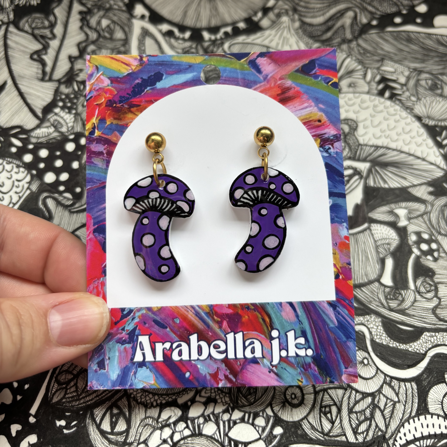 Purple mushroom earrings with white polka dots on a colorful card with 'Arabella j.k.