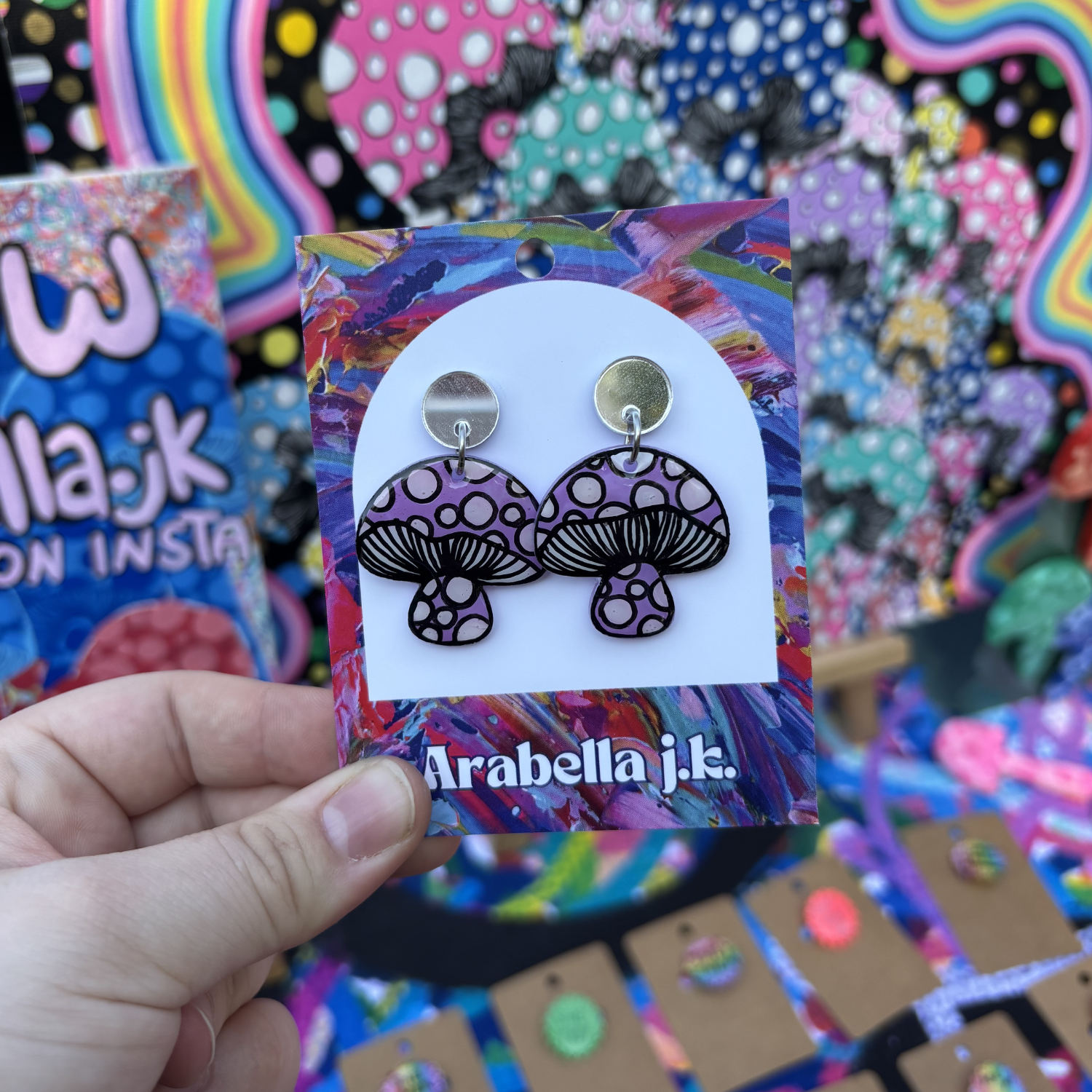 Mushroom-shaped earrings on a colorful 'Arabella j.k.' card held in front of a vibrant background.