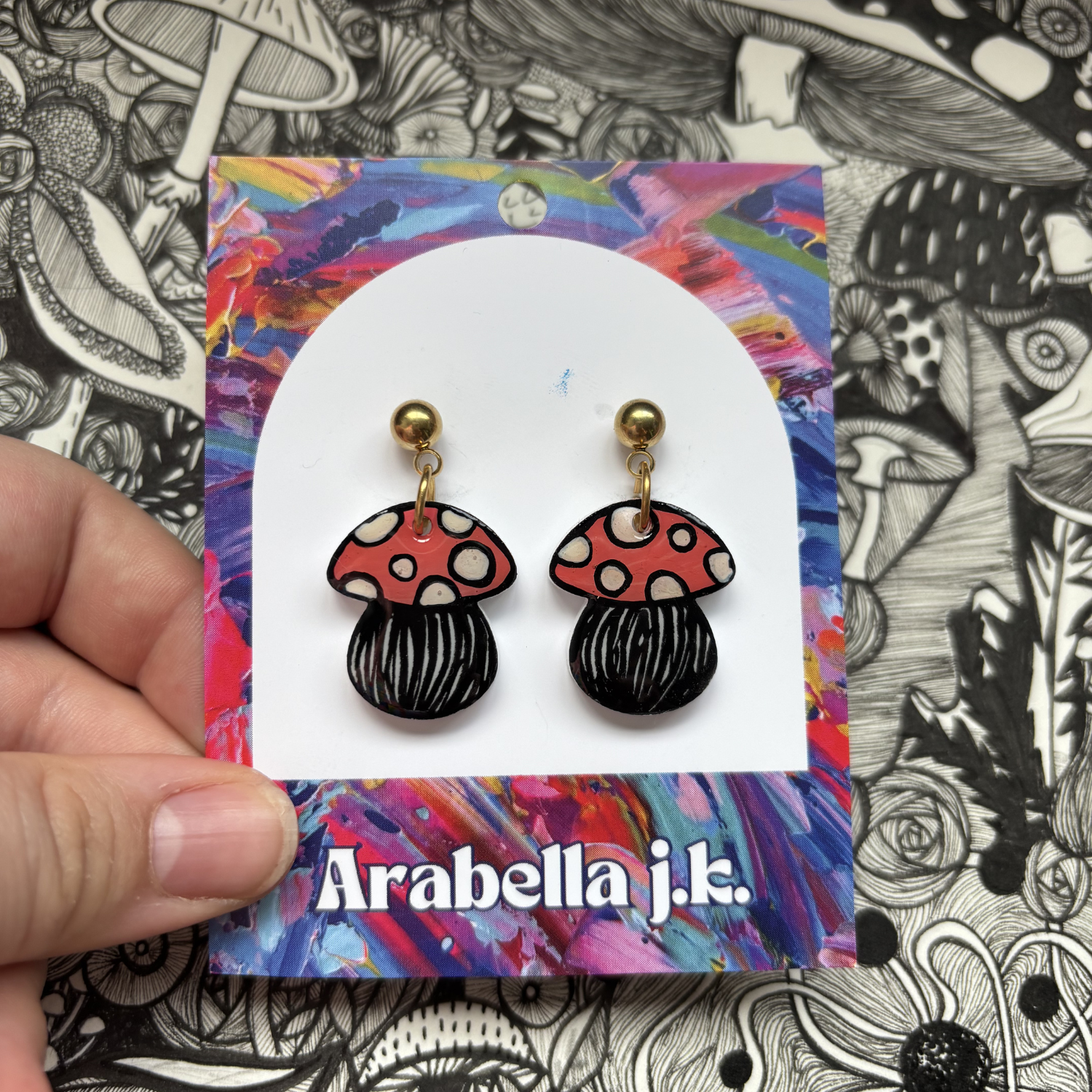 Mushroom-shaped earrings on a colorful 'Arabella j.k.' card against a black and white patterned background.