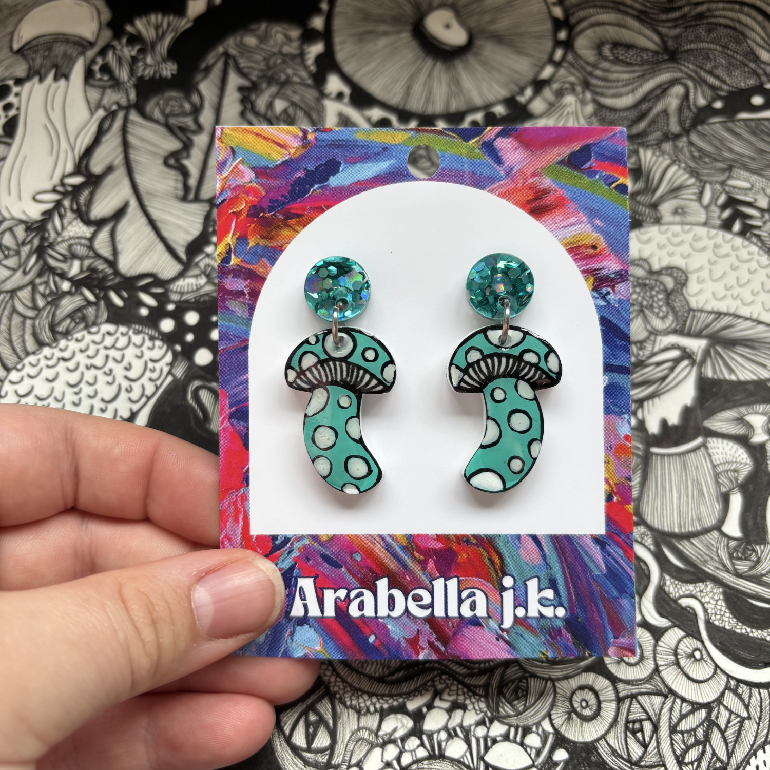 Turquoise mushroom-shaped earrings on a colorful 'Arabella j.k.' card against a black and white patterned background.
