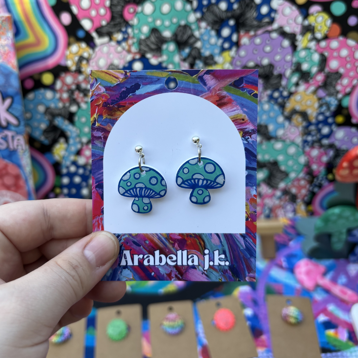 Blue mushroom-shaped earrings on a colorful 'Arabella j.k.' card held by a hand.