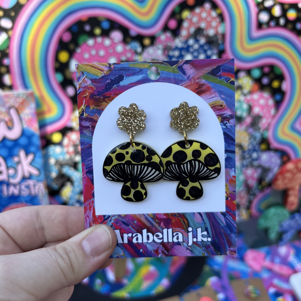 Mushroom-shaped earrings on a colorful 'Arabella j.k.' card against a vibrant, abstract background.